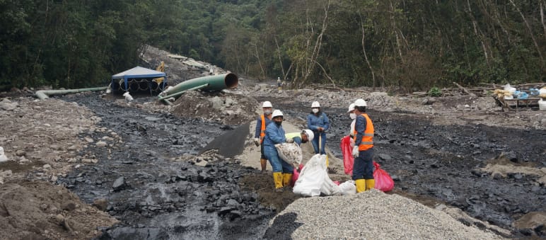 Grupo de petroleiros trabalhando entre dois fluxos de petróleo pesado, que vão se derramando morro abaixo em direção à floresta tropical, na encosta coberta de cascalho. No fundo, vê-se uma parte da tubulação do oleoduto.