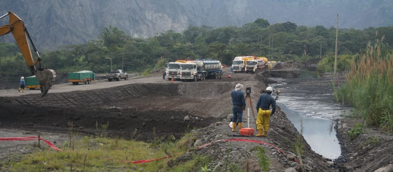 Engenheiros medem a extensão do terreno entre um lago coberto de terra petróleo (à direita) e a visível carregadeira de uma escavadeira cavando mais uma fossa (à esquerda). Ao fundo, estão os caminhões-tanque diante da paisagem montanhosa