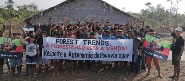 Mais de 30 pessoas do povo Ka'apor estão em pé, diante de uma casa com pilares de madeira e telhado de palha. As pessoas estão com o braço estendido e o punho cerrado. Algumas seguram uma faixa de protesto com a frase: "Forest Trends, a floresta não está