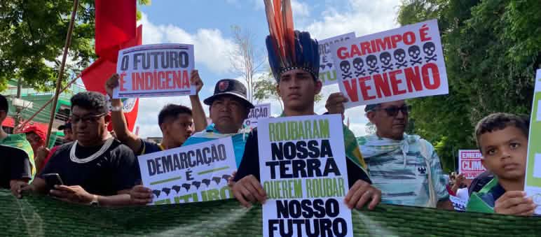 “Marcha dos Povos Indígenas” pela floresta tropical durante a Cúpula da Amazônia em Belém Indígenas marcham em uma fileira e elevam pôsteres com textos como: "Garimpo é veneno" e "O futuro é indígena"