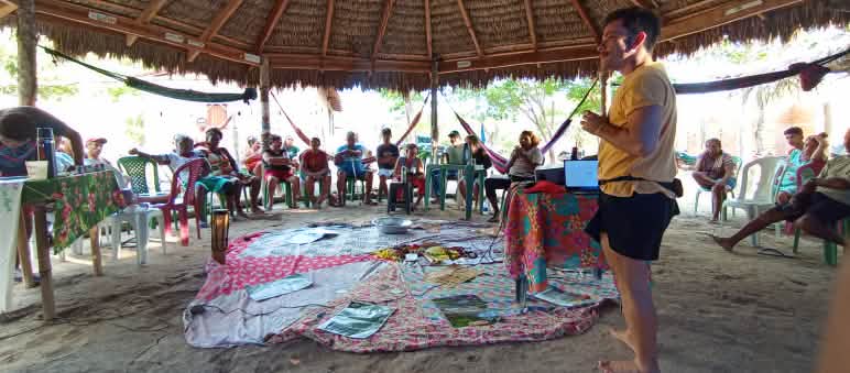 Sentados em círculo em cadeiras, sob uma tenda de madeira e palha, homens e mulheres conversam com um homem que está em pé, ouvindo-os