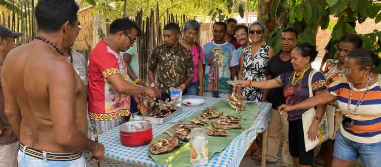 Grupo de 12 pessoas - Mulheres e homens em torno de uma mesa com metades de peixe grelhado.