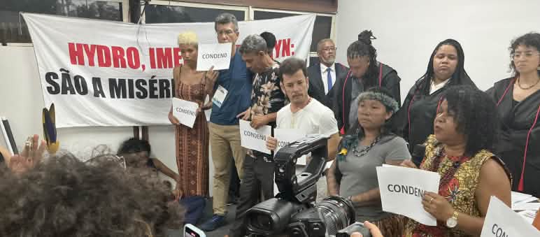 Mais de 10 pessoas estão em pé diante de uma platéia. Algumas pessoas seguram uma placa onde está escrito "condeno", em referência ao veredito do júri