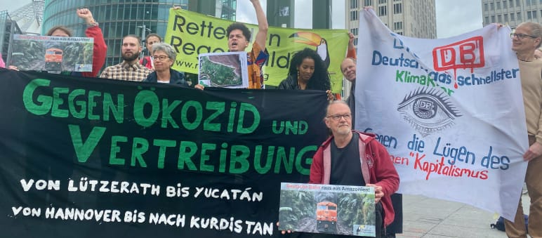 Manifestação na Potsdamer Platz em Berlin, em frente à sede da DB, no fim de maio de 2024 Um grupo de pessoas protesta com grandes faixas coloridas em frente a três torres de escritórios