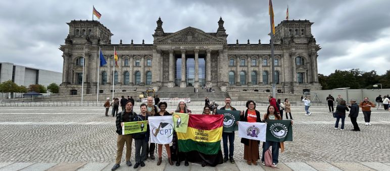 Nove pessoas em pé, segurando bandeiras com logos e nomes de organizações, posam para foto em frente à sede do parlamento alemão