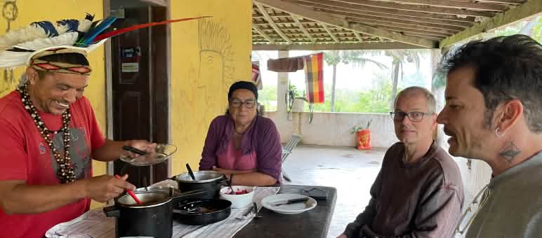 Mãdy Pataxó, adornado com um cocar de penas, sua mãe, Felipe Sabrina e Klaus Schenck, ativistas de Salve a Floresta sentados em torno de uma mesa de madeira preparada para o almoço
