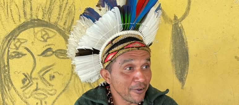 Liderança Mandyn Pataxó usando cocar de penas coloridas em frente a uma parede amarela