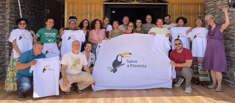 Foto de grupo de 20 pessoas com uma faixa e camisetas com o logo de tucano da associação “Salve a Floresta” (Rettet den Regenwald)