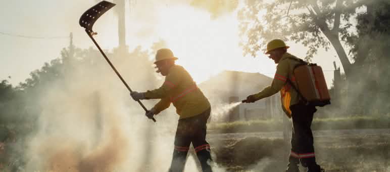 Em meio à fumaça, um homem combate o incêndio com um abafador, enquanto outro utiliza um pulverizador de água costal