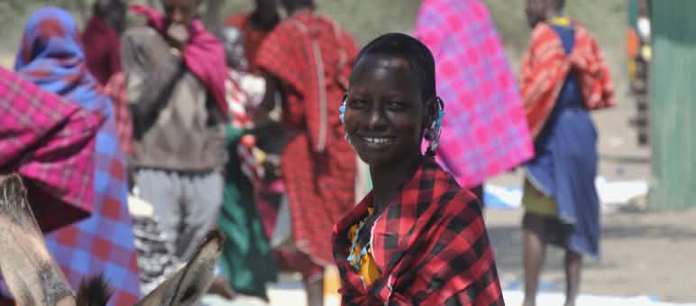 Mulheres e homens do povo Massai com sarongues coloridos em um mercado, uma mulher olha para a câmera.