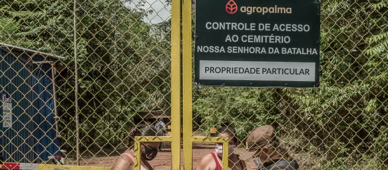 Até mesmo para entrar no seu próprio cemitério, a população local precisa superar obstáculos impostos pela Agropalma, tais como cercas e postos de vigilância Duas mulheres sendo controladas por um guarda por trás de um portão gradeado - Texto inscrito na placa da empresa: Agropalma - Controle de Acesso ao Cemitério Nossa Senhora da Batalha. PROPRIEDADE PARTICULAR