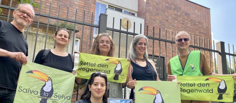 6 pessoas diante dos alambrados do prédio da Embaixada, segurando faixas com as inscrições ”Rettet den Regenwald” e “Salva la Selva” com o tucano-símbolo da associação