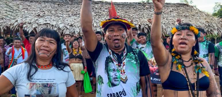 Indígenas Ka'apor protestam contra o projeto planejado de CO₂ em sua floresta tropical Três indígenas Ka'apor - duas mulheres e um homem - com os punhos erguidos em frente a um abrigo coberto com folhas de palmeira