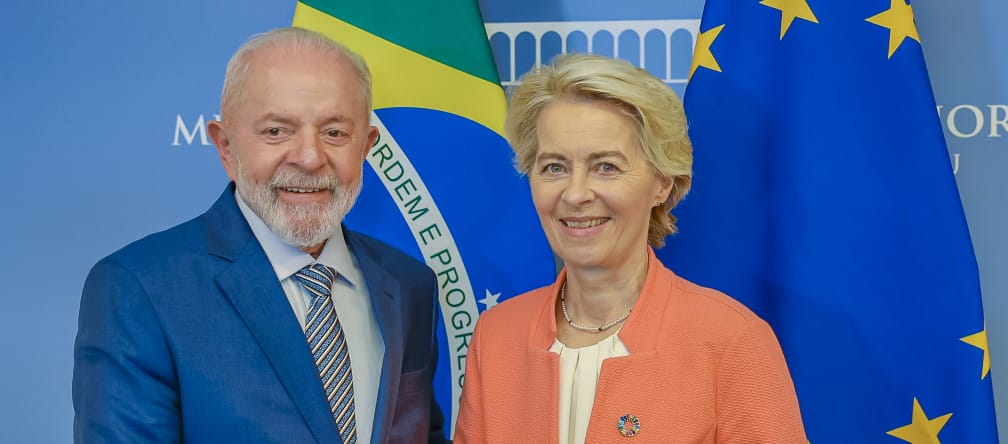 A Presidente da União Europeia, Ursula von der Leyen e o Presidente brasileiro, Lula, apertam-se as mãos diante das bandeiras nacionais