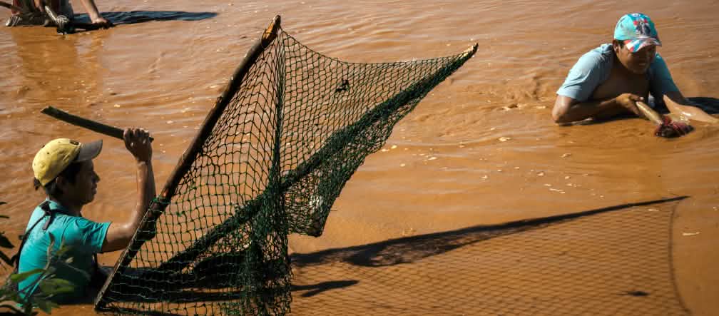 Três homens pescando, em pé, com água até a cintura, em um rio marrom
