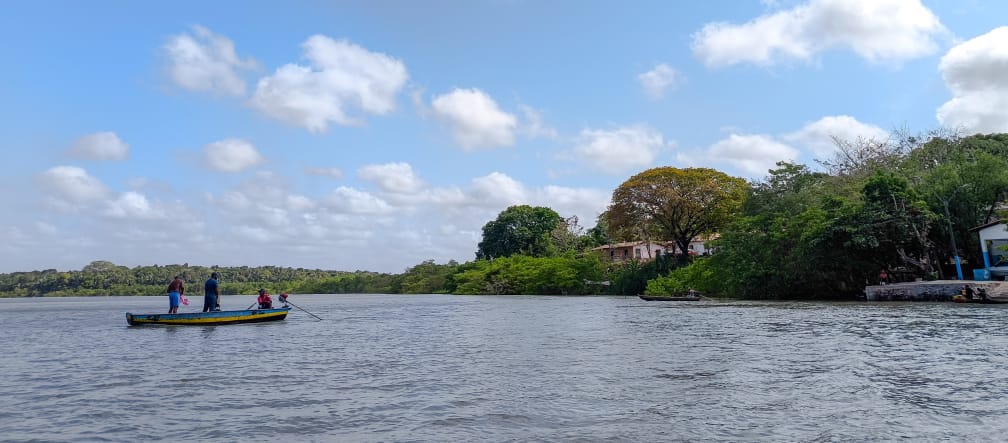 Sob o céu azul, um pequeno barco navega pelo mar com três pescadores. À direita estão pequenas casas no meio de uma floresta.