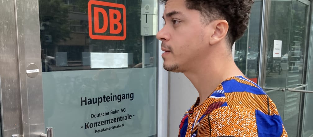 Um jovem está parado com um envelope em frente a uma porta de vidro com a etiqueta DB Entrada principal da sede do Grupo