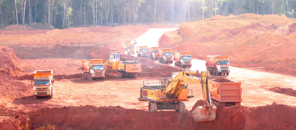 Uma escavadeira empurra o solo vermelho de um poço para dentro de um caminhão. Atrás dela, uma fila de caminhões de espera e máquinas de construção é visível. Árvores na floresta tropical em segundo plano