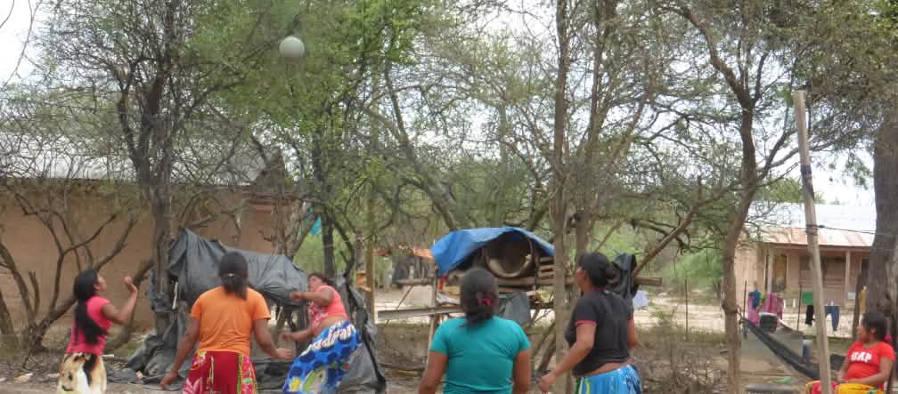 Cinco mulheres indígenas jogando bola entre árvores e casas de madeira