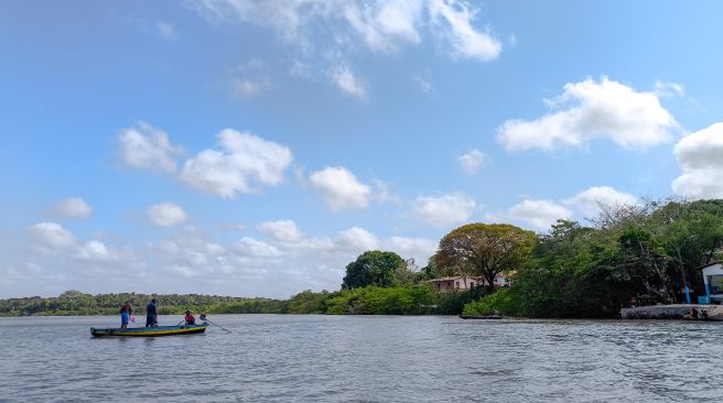 Sob o céu azul, um pequeno barco navega pelo mar com três pescadores. À direita estão pequenas casas no meio de uma floresta.