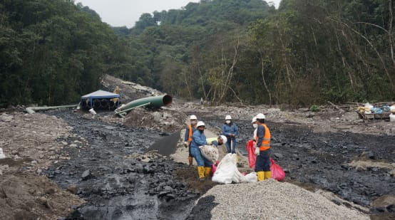 Grupo de petroleiros trabalhando entre dois fluxos de petróleo pesado, que vão se derramando morro abaixo em direção à floresta tropical, na encosta coberta de cascalho. No fundo, vê-se uma parte da tubulação do oleoduto.