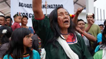 Moradores nativos, mulheres, crianças e homens diante de uma faixa, fazendo uma manifestação