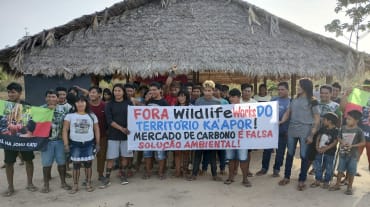 Mais de 30 pessoas do povo Ka'apor estão em pé, diante de uma casa com pilares de madeira e telhado de palha. As pessoas seguram uma faixa de protesto com a frase: "Fora Wildlife Works do território Ka'apor! Mercado de carbono é falsa solução ambiental!"