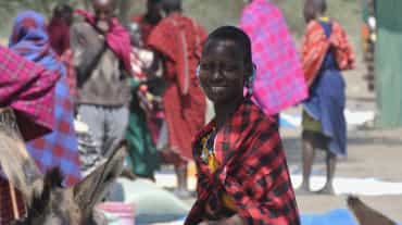 Mulheres e homens do povo Massai com sarongues coloridos em um mercado, uma mulher olha para a câmera.