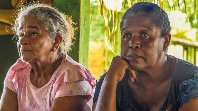 Dica e Natividade Akroá Gamella durante roda de conversa na aldeia Tabarelzinho Duas mulheres indígenas sentadas lado a lado, observando algo. A da esquerda veste blusa rosa; a da direita veste blusa azul escuro e apoia o rosto sobre a mão direita.