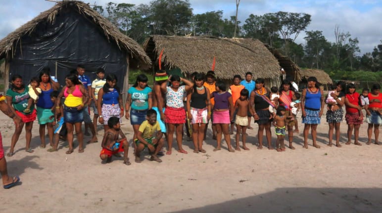 Pessoas da etnia Ka’apor em fila dupla diante de duas cabanas na floresta