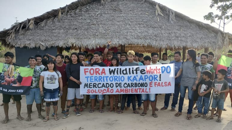 Membros do povo Ka'apor fazem protesto contra a presença de empresa norte-americana causando conflitos em seu território Mais de 30 pessoas do povo Ka'apor estão em pé, diante de uma casa com pilares de madeira e telhado de palha. As pessoas seguram uma faixa de protesto com a frase: "Fora Wildlife Works do território Ka'apor! Mercado de carbono é falsa solução ambiental!"
