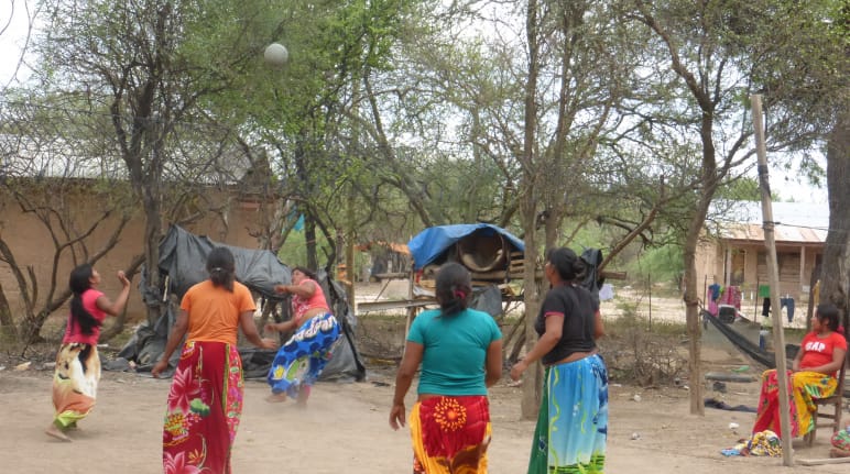 Cinco mulheres indígenas jogando bola entre árvores e casas de madeira