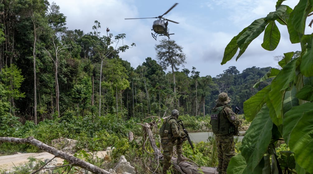 Dois agentes do IBAMA, armados e vestidos de uniformes de camuflagem, sob um helicóptero sobrevoando área desmatada na floresta