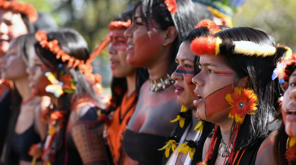 Oito mulheres indígenas, lado a lado, com enfeites de plumas e penas em suas cabeças, brincos de penas amarelas e vermelhas, e pintura vermelha e preta no rosto