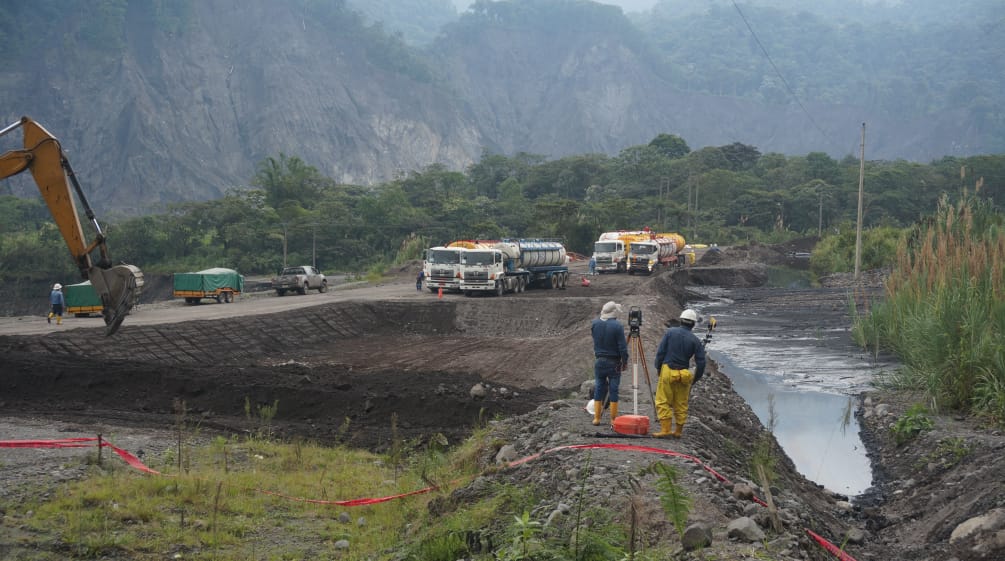 Engenheiros medem a extensão do terreno entre um lago coberto de terra petróleo (à direita) e a visível carregadeira de uma escavadeira cavando mais uma fossa (à esquerda). Ao fundo, estão os caminhões-tanque diante da paisagem montanhosa