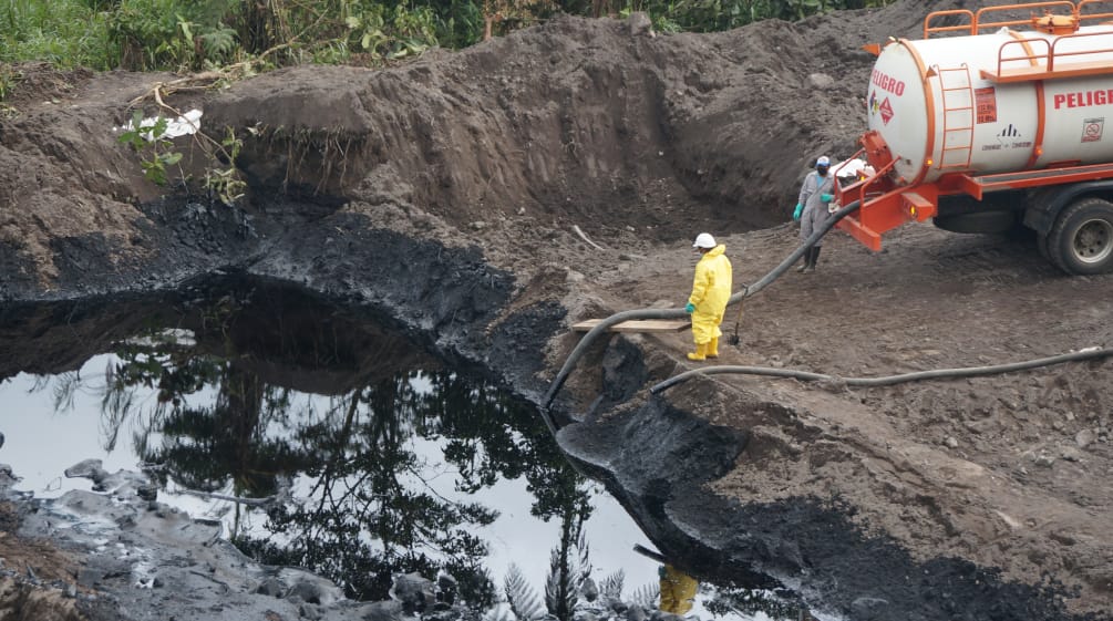 Dois petroleiros transferem petróleo com uma mangueira de um lago coberto de terra para um caminhão-tanque.