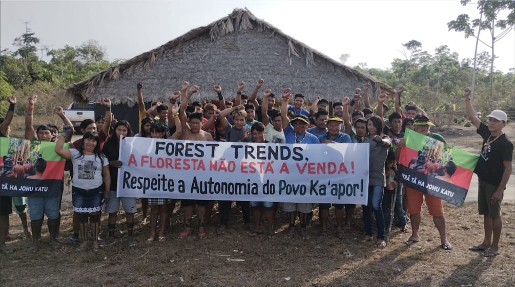 Mais de 30 pessoas do povo Ka'apor estão em pé, diante de uma casa com pilares de madeira e telhado de palha. As pessoas estão com o braço estendido e o punho cerrado. Algumas seguram uma faixa de protesto com a frase: "Forest Trends, a floresta não está