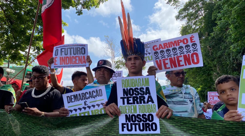 Indígenas marcham em uma fileira e elevam pôsteres com textos como: "Garimpo é veneno" e "O futuro é indígena"
