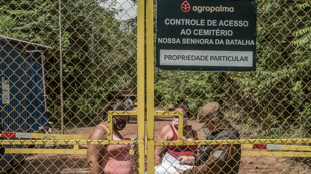 Duas mulheres sendo controladas por um guarda por trás de um portão gradeado - Texto inscrito na placa da empresa: Agropalma - Controle de Acesso ao Cemitério Nossa Senhora da Batalha. PROPRIEDADE PARTICULAR